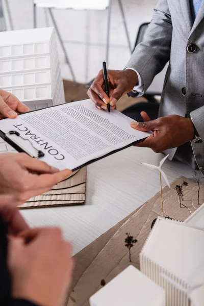 Teilansicht eines afrikanisch-amerikanischen Geschäftsmannes bei Vertragsunterzeichnung in der Nähe von Ökostromkraftwerk-Modell und Geschäftspartnern im verschwommenen Vordergrund — Stockfoto