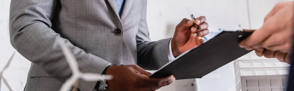 Vista parcial del empresario sosteniendo portapapeles cerca de socio afroamericano y modelo de generador eólico en primer plano borroso, pancarta - foto de stock