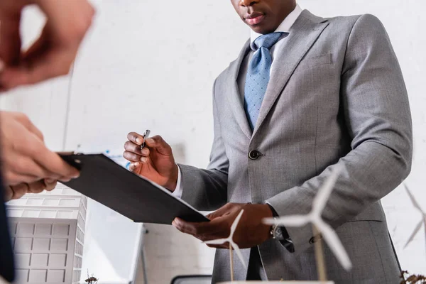 Partial view of african american businessman holding pen near clipboard and business partner on blurred foreground — Stock Photo