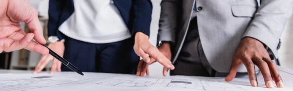Cropped view of multiethnic business partners pointing at blueprint during meeting, banner — Stock Photo