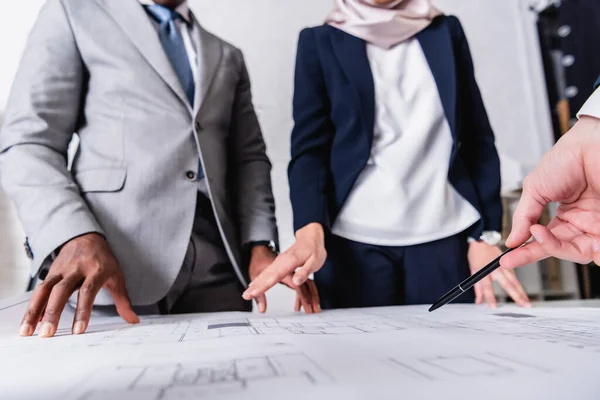 Cropped view of businessman pointing with pen at blueprint near interracial business partners on blurred background — Stock Photo