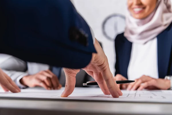 Abgeschnittene Ansicht einer lächelnden arabischen Frau in der Nähe von Blaupausen und multikulturellen Geschäftsleuten, verschwommener Hintergrund — Stockfoto