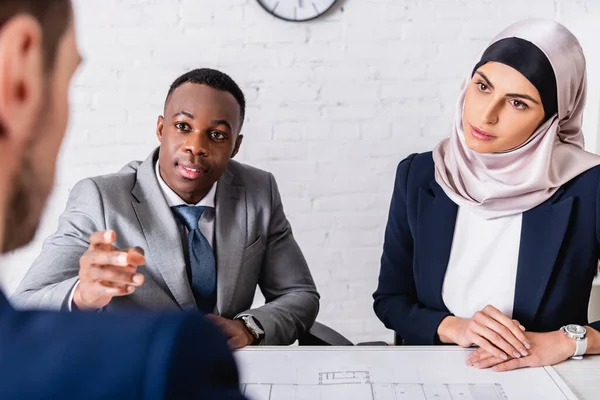 Uomo d'affari afroamericano che punta con mano vicino al progetto e partner commerciali multietnici, primo piano sfocato — Foto stock