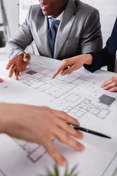 Partial view of businesswoman pointing at blueprint during meeting with multicultural business partners, blurred foreground — Stock Photo