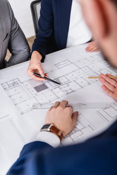 Cropped view of business partners pointing with pencil and pen at blueprint, blurred foreground — Stock Photo