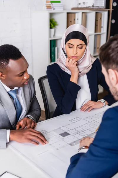 Serious multiethnic business partners looking at blueprint near interpreter, blurred foreground — Stock Photo