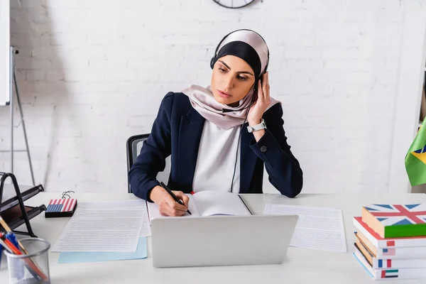 Arabian interpreter in headset writing in notebook near laptop, digital translator and dictionaries of foreign languages — Stock Photo