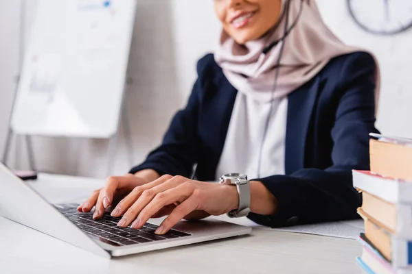 Cropped view of smiling arabian translator typing on laptop near dictionaries of foreign languages, blurred background — Stock Photo