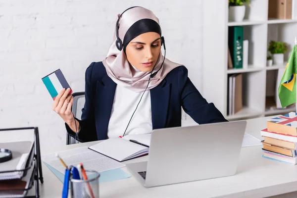 Arabisch-Dolmetscher im Kopfhörer hält digitalen Übersetzer während der Arbeit am Laptop in der Nähe von Wörterbüchern, verschwommener Vordergrund — Stockfoto