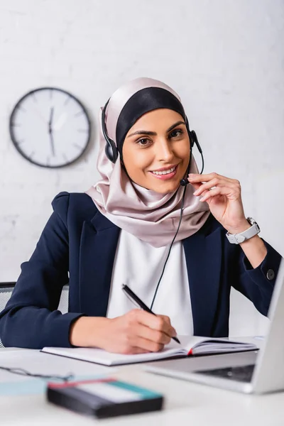 Intérprete árabe feliz em headset segurando caneta perto notebook e tradutor digital com uae bandeira emblema em primeiro plano borrado — Fotografia de Stock