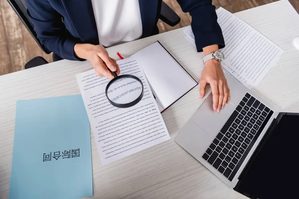 Partial view of interpreter holding magnifier near documents and paper with chinese hieroglyphs. Translation: 