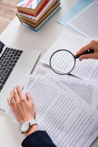 Partial view of translator holding magnifier above documents near laptop and dictionaries of foreign languages — Stock Photo