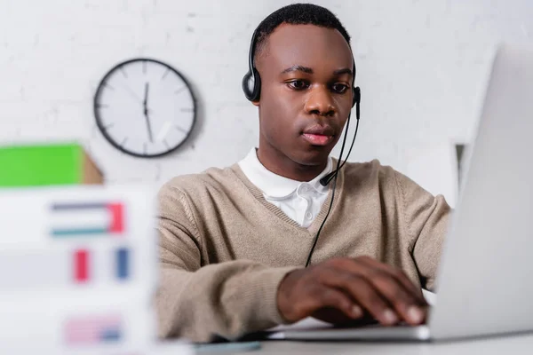 Young african american translator in headset working at laptop on blurred foreground — Stock Photo