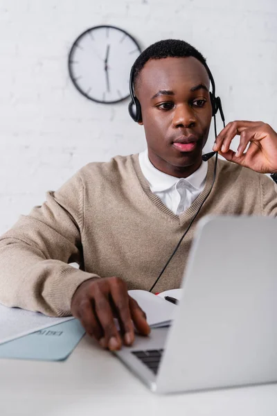 Jovem tradutor afro-americano em fone de ouvido trabalhando no laptop em primeiro plano borrado — Fotografia de Stock