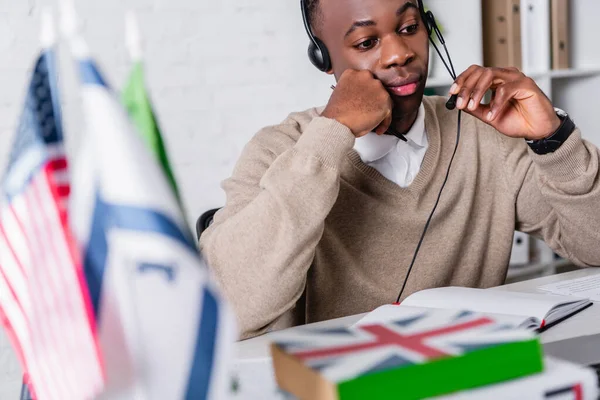 Thoughtful african american translator in headset working near international flags and dictionaries on blurred foreground — Stock Photo