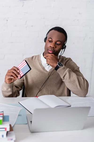 Thoughtful african american interpreter in headset holding digital translator with usa flag emblem near notebook and laptop on blurred foreground — Stock Photo