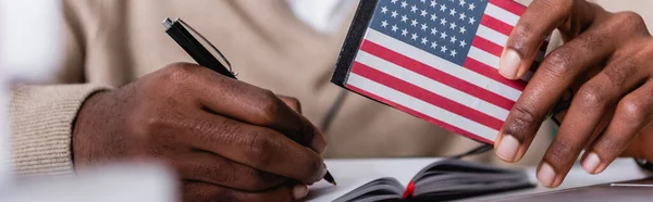 Messa a fuoco selettiva dell'interprete afroamericano che scrive nel taccuino mentre tiene il traduttore digitale con l'emblema della bandiera degli Stati Uniti, la vista ritagliata, banner — Foto stock