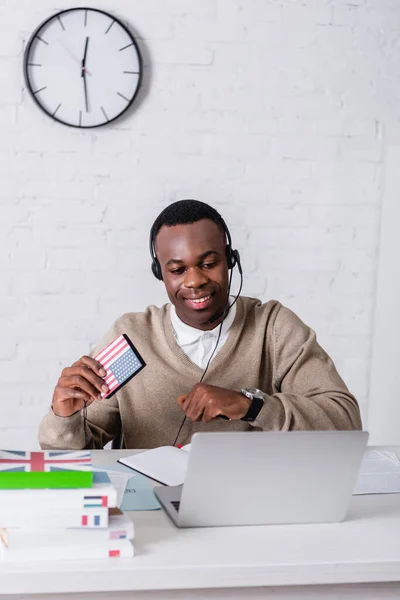 Cheerful african american interpreter holding digital translator with usa flag emblem near laptop and dictionaries on blurred foreground — Stock Photo