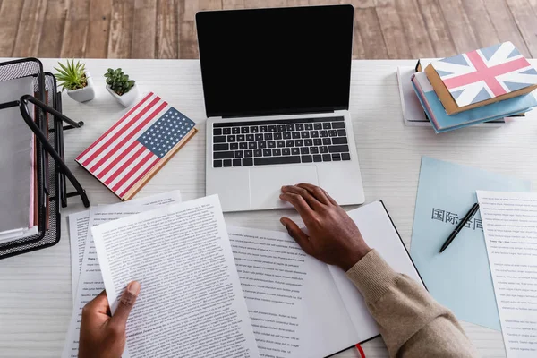 Vue partielle du traducteur afro-américain travaillant avec des documents près d'un ordinateur portable et des dictionnaires avec des Etats-Unis et de grands drapeaux britanniques sur les couvertures — Photo de stock