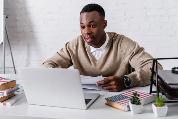 Amazed african american translator working at laptop near dictionaries on blurred foreground — Stock Photo