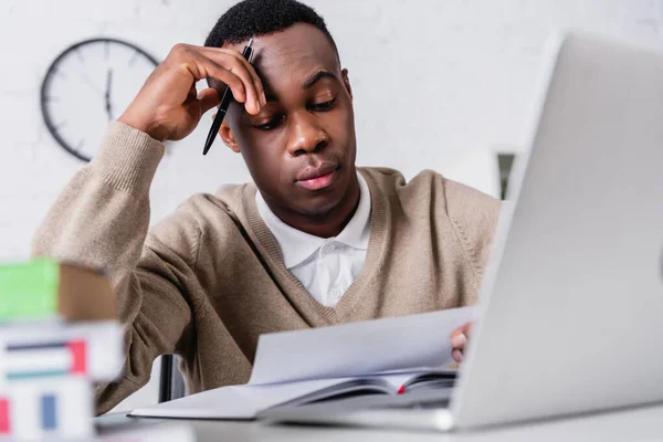 Thoughtful afican american translator holding pen while working with document near laptop on blurred foreground — Stock Photo