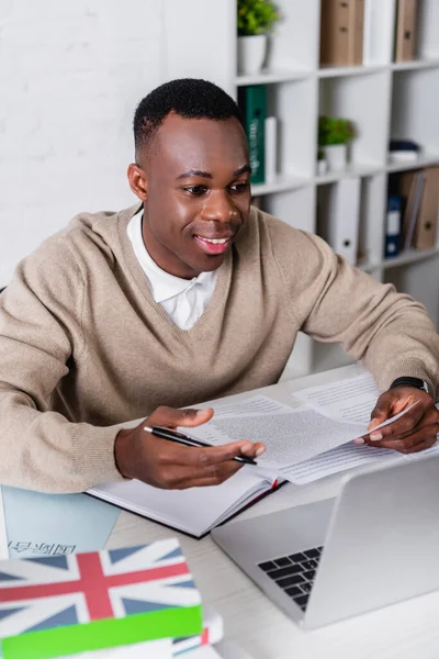 Sorridente tradutor afro-americano trabalhando com documentos perto de laptop e dicionários em primeiro plano borrado — Fotografia de Stock