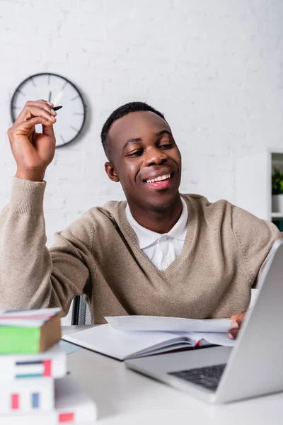 Cheerful african american translator working with document near laptop and dictionaries on blurred foreground — Stock Photo