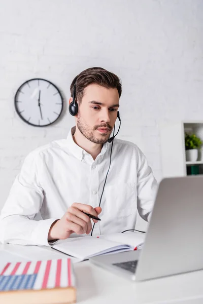 Young translator in headset holding pen near notebook and dictionary on blurred foreground — стоковое фото