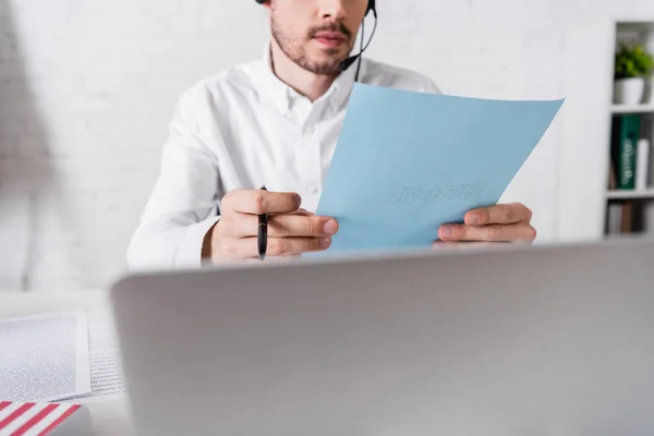 Partial view of translator in headset working with document near laptop on blurred foreground — Stock Photo