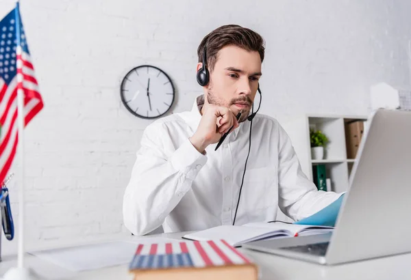 Traduttore premuroso in auricolare che lavora vicino a laptop, bandiera americana e dizionario in primo piano sfocato — Foto stock