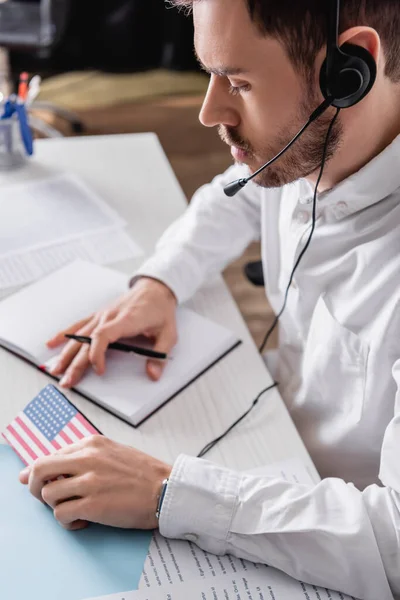 Jovem intérprete em headset segurando caneta e tradutor digital com EUA bandeira emblema, fundo borrado — Fotografia de Stock