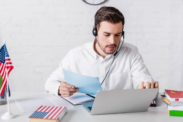 Junger Übersetzer im Headset mit Dokument, während er am Laptop in der Nähe von Wörterbüchern und amerikanischer Flagge arbeitet — Stockfoto