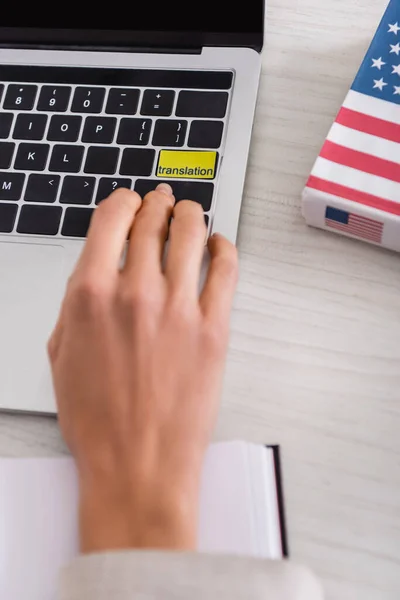 Vista parcial del intérprete presionando la tecla de traducción en el ordenador portátil cerca del diccionario inglés americano, primer plano borroso - foto de stock