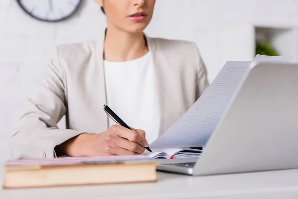 Cropped view of translator writing in notebook while holding document near laptop, blurred foreground — Stock Photo