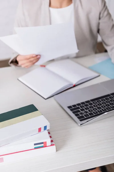 Cropped view of translator holding documents near blank notebook, laptop, and dictionaries, blurred background — Stock Photo