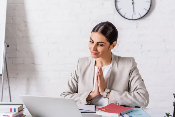 Smiling translator showing greeting gesture during video call on laptop near dictionaries — Stock Photo