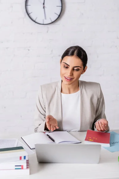 Smiling translator pointing with hand during video call near notebook and dictionaries on blurred foreground — Stock Photo