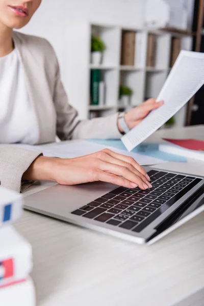 Partial view of translator holding documents while typing on laptop, blurred foreground — Stock Photo