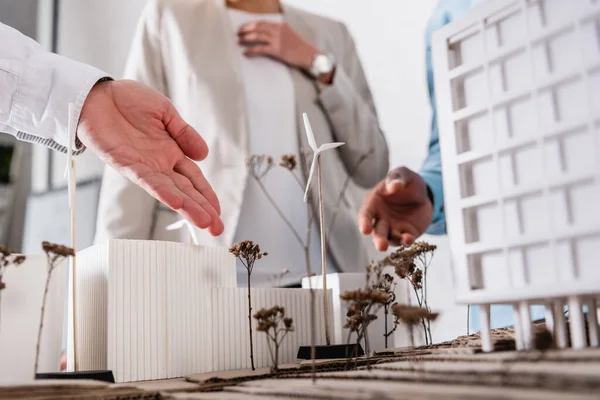 Ponto de vista do empresário que aponta para uma central de energia alternativa próxima do modelo de construção e para parceiros multiétnicos de fundo turvo — Fotografia de Stock