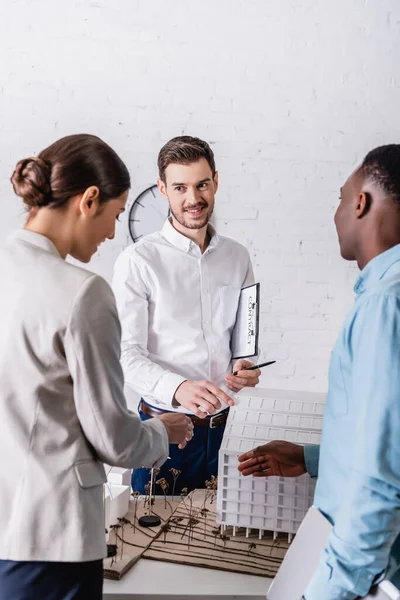 Glücklicher Übersetzer, der mit dem Finger auf den Aufbau von Modellen in der Nähe von interrassischen Geschäftspartnern zeigt — Stockfoto