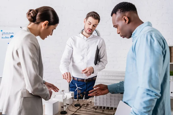 Multiethnic business partners pointing at model of green power station in office — Stock Photo