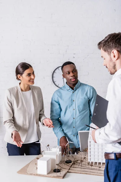 Donna d'affari sorridente che indica il modello di centrale energetica verde vicino a partner commerciali multietnici — Foto stock