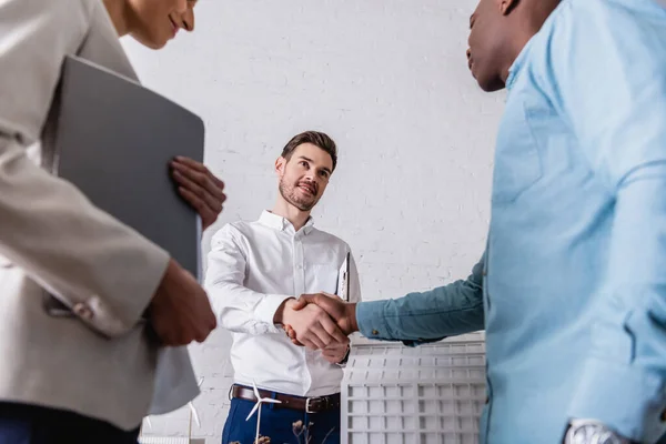 Empresário afro-americano apertando as mãos com intérprete perto de empresária e modelo de construção, foreground borrado — Fotografia de Stock