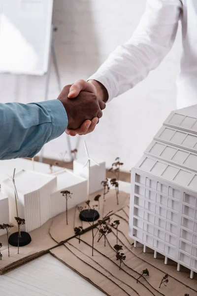 Cropped view of interracial business partners shaking hands near models of building and alternative power station — Stock Photo