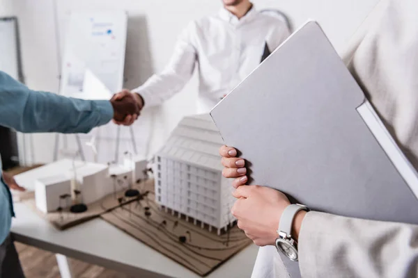 Cropped view of translator with paper folder, and interracial partners shaking hands near green energy station model, blurred background — Stock Photo