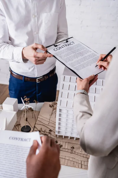 Vista ritagliata della donna d'affari che firma un contratto vicino ai partner commerciali interrazziali e alla stazione energetica alternativa, primo piano sfocato — Foto stock