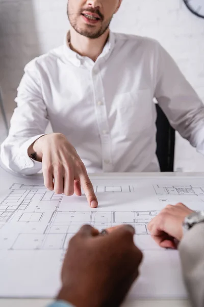 Abgeschnittene Ansicht eines Geschäftsmannes, der mit dem Finger auf Blaupause in der Nähe multiethnischer Geschäftspartner zeigt, auf verschwommenem Vordergrund — Stockfoto