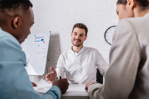 Smiling businessman pointing with hand near blueprint and interracial business partners on blurred foreground — Stock Photo