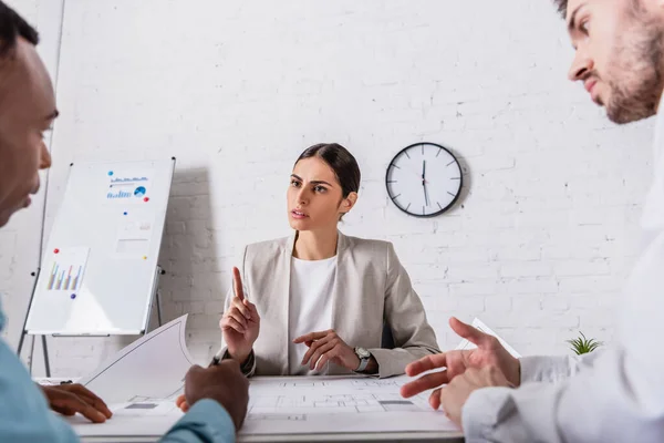 Serious businesswoman showing attention gesture during meeting with multicultural business partners near blueprint, blurred foreground — Stock Photo