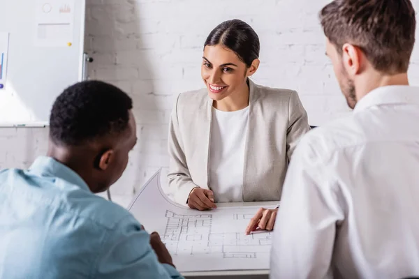 Lächelnde Geschäftsfrau zeigt mit dem Finger auf Blaupause in der Nähe von interrassischen Geschäftspartnern auf verschwommenem Vordergrund — Stockfoto